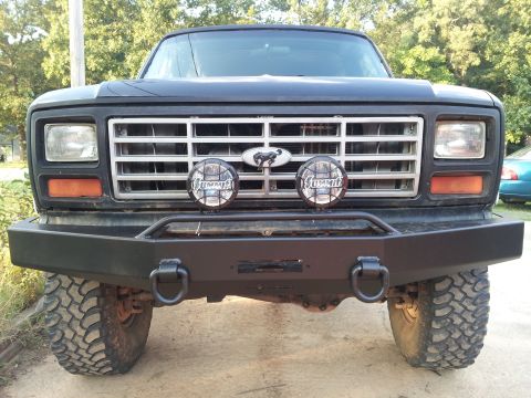 1986 Ford Bronco - This black Ford Bronco features an aggressive aftermarket bumper and auxiliary lights, with the iconic Ford badge prominently displayed on…