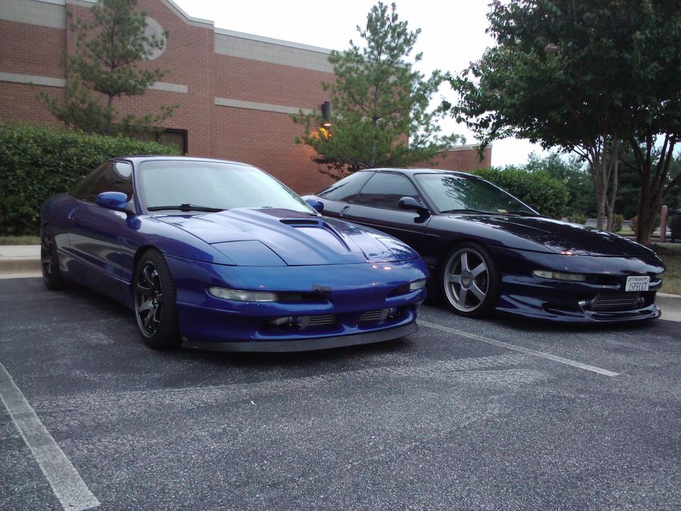 Two 1993 Ford Probes parked side-by-side in a parking lot. The front car is blue, the rear car is dark blue