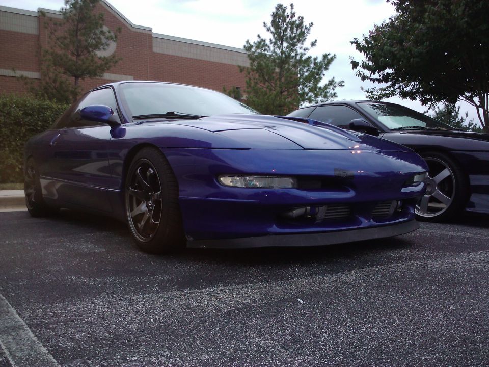 Blue sports coupe parked next to another car. Has aftermarket wheels and a front lip