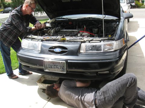 1992 Ford Taurus - Two people are working on the engine of a black Ford Taurus, with one person lying on the ground to access the underside.