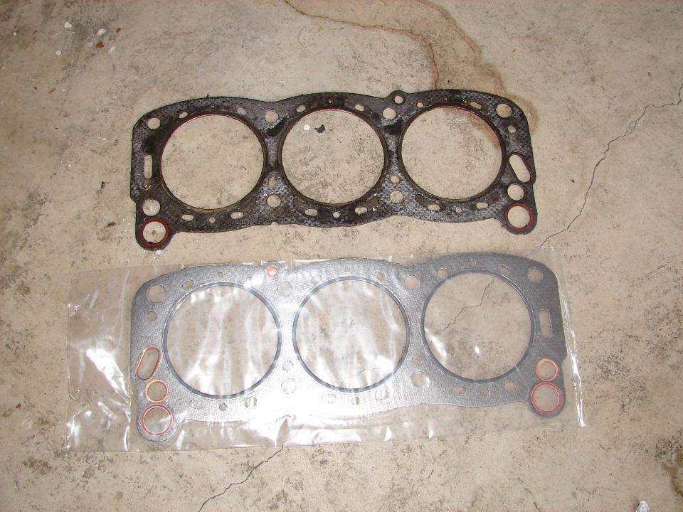 Two engine gaskets on a concrete floor. One is used, the other is new in plastic