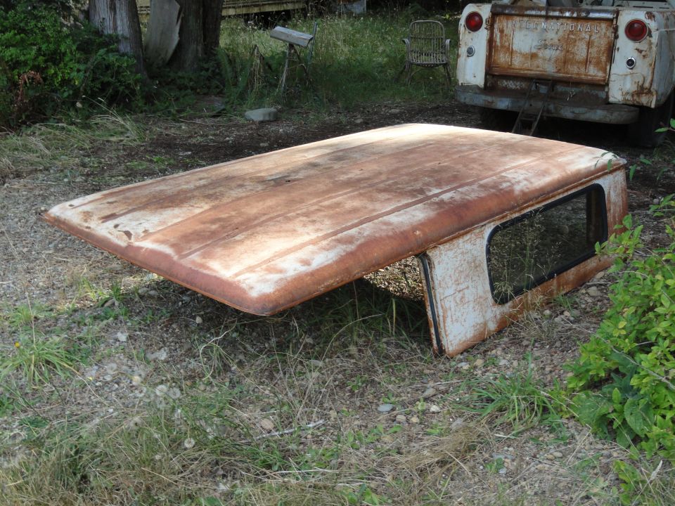 Rusted truck cap with a window, lying on the ground. A white pickup truck is visible in the background