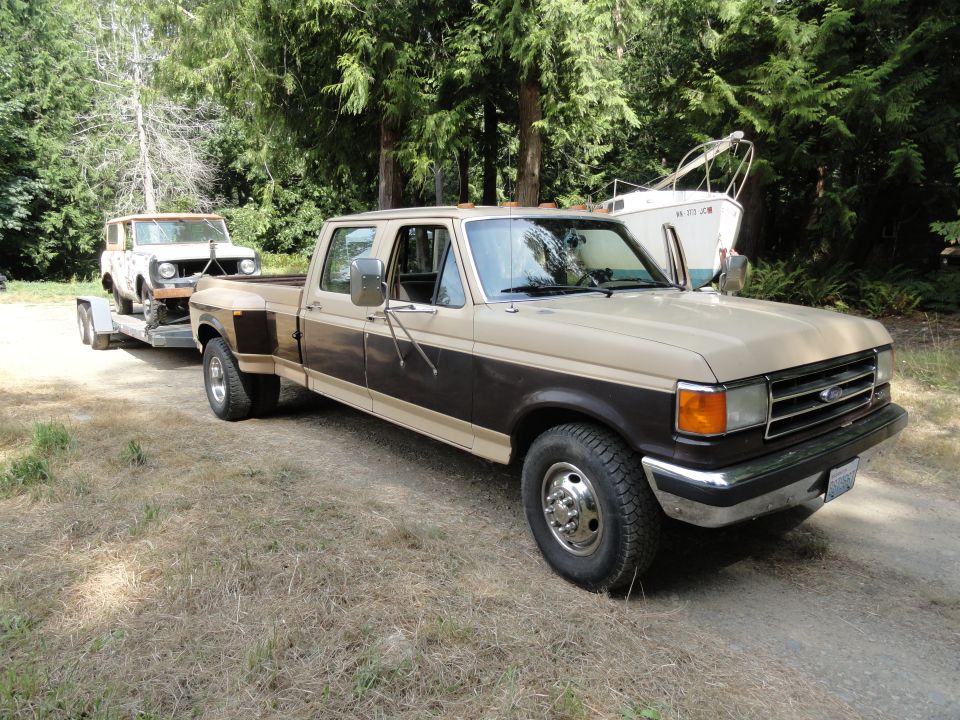 Ford F-350 dually pickup truck towing a white International Scout on a trailer. The truck is tan and dark brown. Trees and a boat are in the background