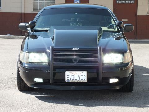 2003 Ford Crown Victoria - Headlights blaze on a black Crown Victoria with a brush guard parked near a Custom Mfg Co. wall.