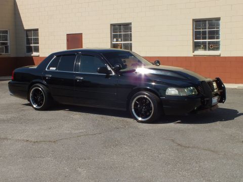 2003 Ford Crown Victoria - Sun glares off a black sedan's roof at a front-quarter angle with brush guard and lip wheels.