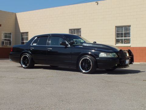 2003 Ford Crown Victoria - Black sedan with a brush guard and chrome-lip wheels parked at a front angle by a tan wall.