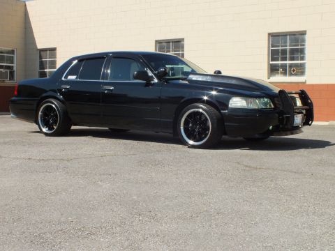 2003 Ford Crown Victoria - Lowered black sedan with brush guard and chrome-lip wheels angled toward the camera in a lot.