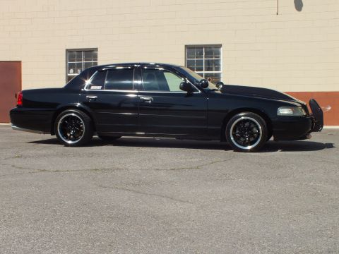 2003 Ford Crown Victoria - Side profile of a lowered black Crown Victoria sedan with black multi-spoke wheels parked beside a tan masonry building.