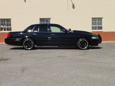2003 Ford Crown Victoria - Passenger-side view of a black Crown Victoria sedan dropped over black wheels in front of a tan brick wall.