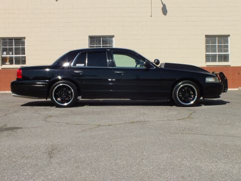 2003 Ford Crown Victoria - Driver-side profile of a lowered black Crown Victoria with red taillights and black wheels by a tan building.