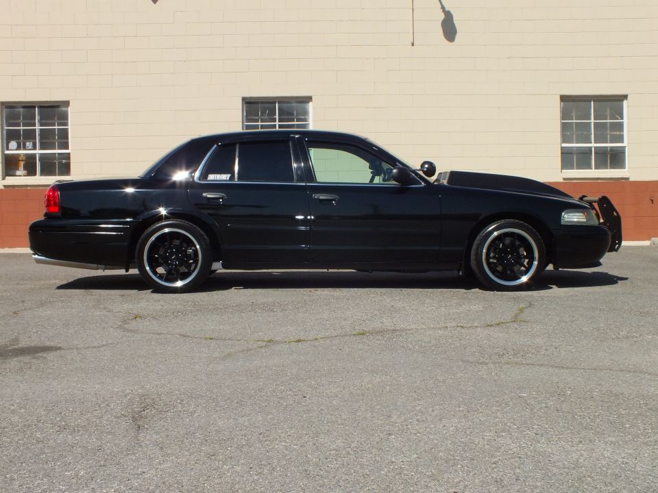 Black Crown Victoria driver-side profile, red taillight, black multi-spoke wheels, tan building
