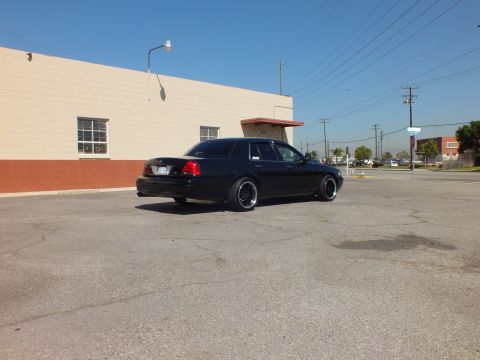 2003 Ford Crown Victoria - Distant rear three-quarter of a black Crown Victoria sedan on a wide asphalt lot under a clear sky.