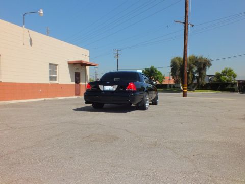 2003 Ford Crown Victoria - Black Crown Victoria sedan seen from rear at distance across a sunlit asphalt lot.