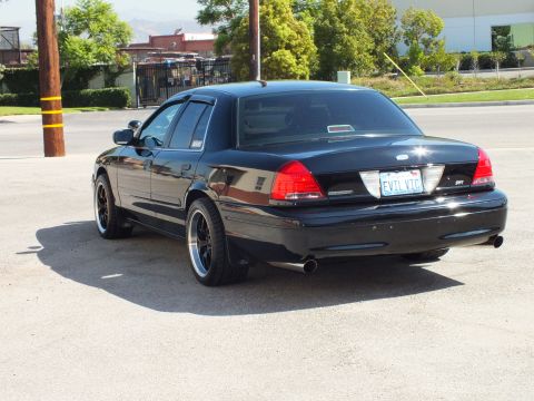 2003 Ford Crown Victoria - Driver-rear three-quarter of a black Crown Victoria on black wheels beside a gated industrial lot.