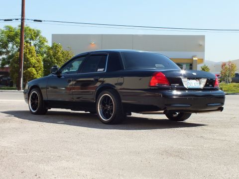 2003 Ford Crown Victoria - Driver-side rear three-quarter of a lowered black Crown Victoria on black wheels by a white building.