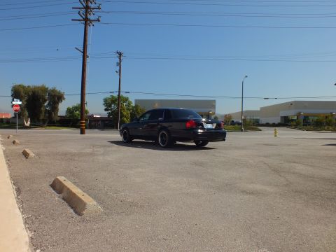 2003 Ford Crown Victoria - Distant rear three-quarter view of a black full-size sedan on black wheels in a wide commercial parking lot.