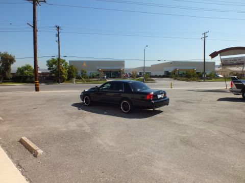 2003 Ford Crown Victoria - Lowered black sedan on black rims sits in a vast parking lot with industrial buildings behind.