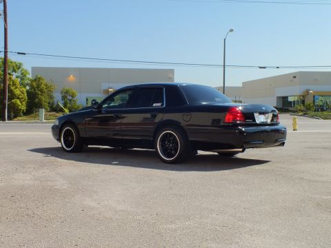 2003 Ford Crown Victoria - Rear quarter of a black sedan showing dark tail lights and black mesh wheels with whitewalls.