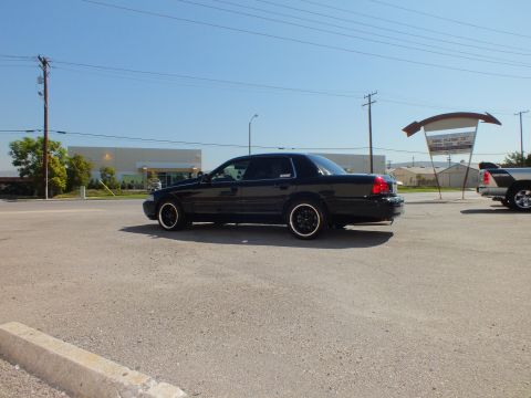 2003 Ford Crown Victoria - Driver-side rear view of a black full-size sedan parked across a wide commercial lot.