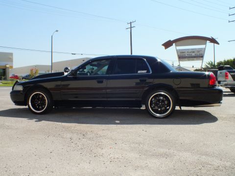 2003 Ford Crown Victoria - Side view of a black four-door sedan, tinted windows, parked before a metal industrial building.