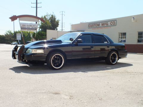 2003 Ford Crown Victoria - Black full-size sedan with grille guard and black rims, parked outside an industrial shop.