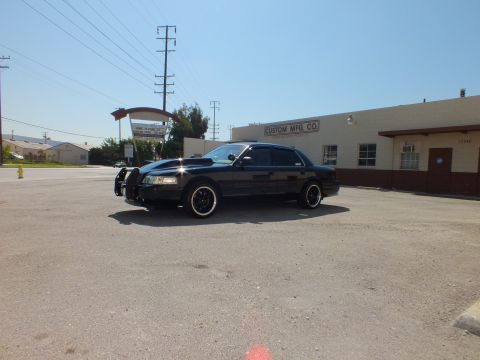 2003 Ford Crown Victoria - Black sedan with push bar viewed from front quarter in a wide industrial parking lot.