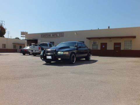 2003 Ford Crown Victoria - Black sedan with grille guard sits in a lot beside a pickup with the shop building behind.