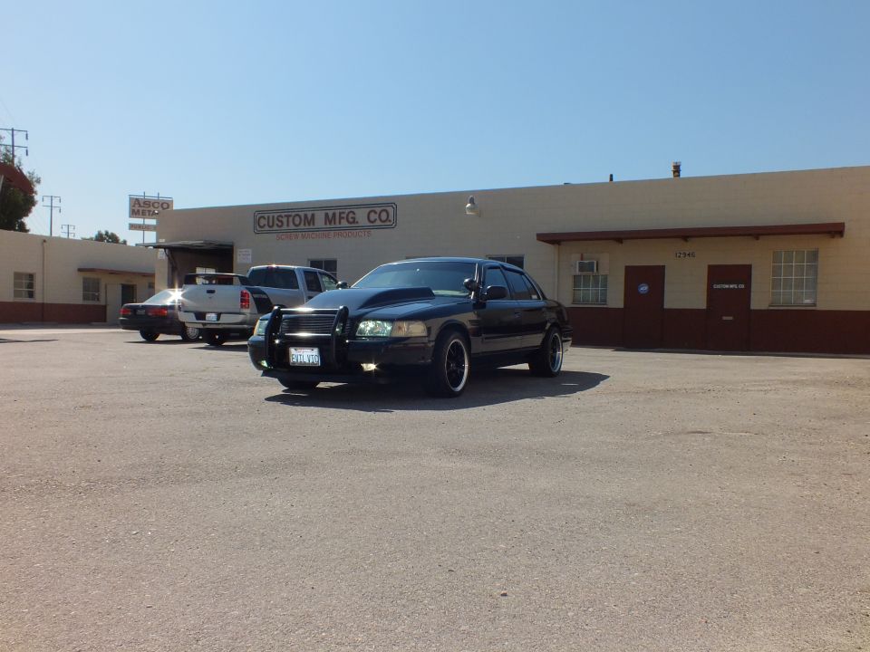 Black Crown Victoria with push bar parked before Custom Mfg Co industrial building