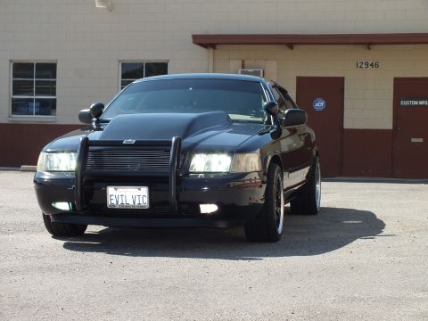 2003 Ford Crown Victoria - Head-on look at a black sedan with grille guard and EVIL VIC plate outside an industrial shop.