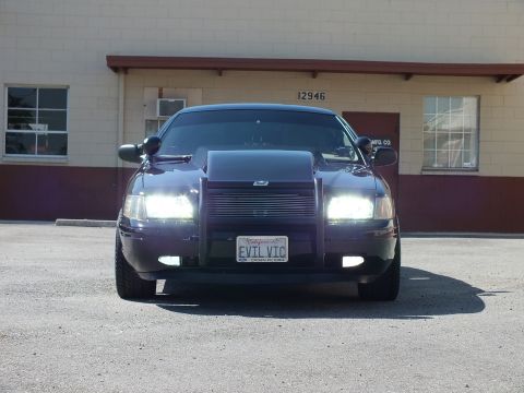 2003 Ford Crown Victoria - Front view of a black sedan with lit headlights, grille guard and a California EVIL VIC plate.