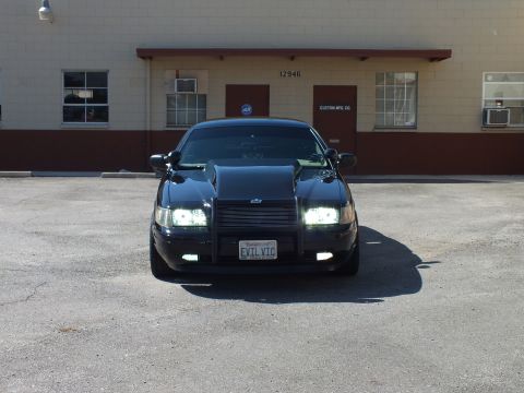 2003 Ford Crown Victoria - Black sedan viewed head-on with lit headlights, push bar and EVIL VIC vanity plate.