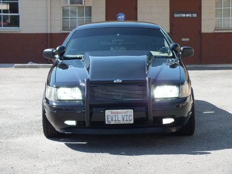 2003 Ford Crown Victoria - Front of a black sedan with grille guard, lit headlights and a California EVIL VIC plate.