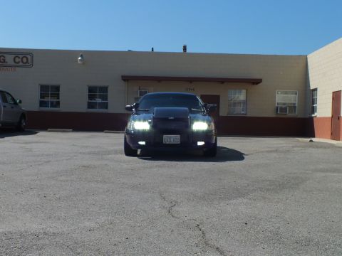 2003 Ford Crown Victoria - Black sedan with headlights lit sits across a wide lot fronting a tan industrial building.