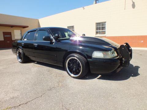 2003 Ford Crown Victoria - Side profile of the black full-size sedan on gloss-black wheels, parked beside a beige building.