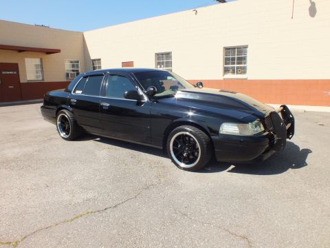 2003 Ford Crown Victoria - Front three-quarter shot of the lowered black sedan with chrome push bar on cracked pavement.