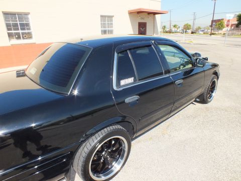 2003 Ford Crown Victoria - Rear three-quarter of the black full-size sedan, heavy window tint and gloss-black wheels.