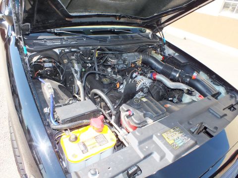 2003 Ford Crown Victoria - Open engine compartment showing a V8, yellow-top battery and air intake on a dark sedan outdoors.