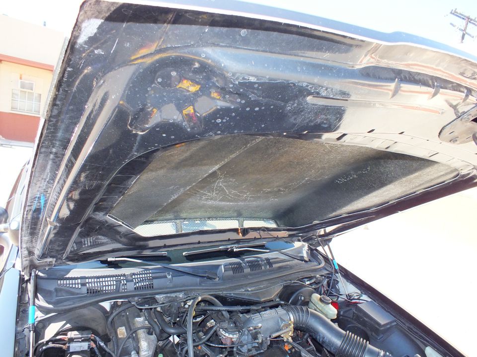 Underside of open black car hood with worn insulation pad and scattered debris, engine bay below