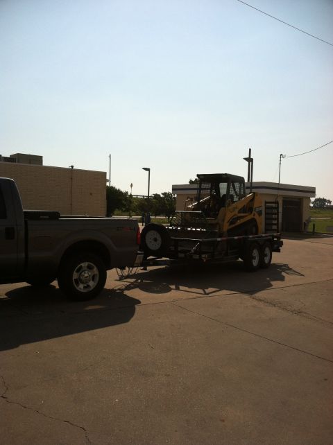 1986 Ford Bronco - A pickup truck tows a trailer carrying a skid steer loader on a sunny day.