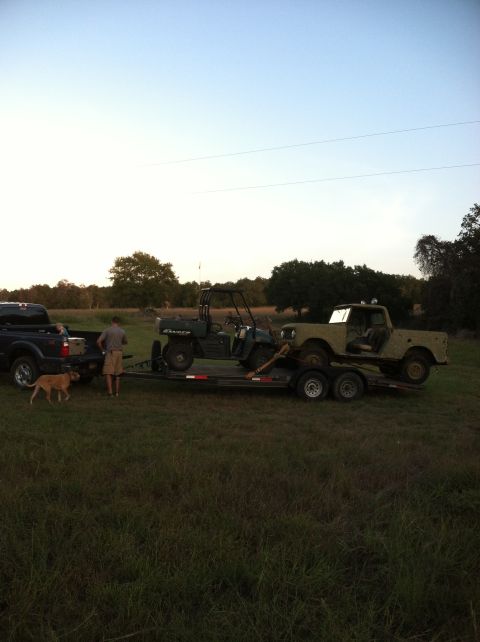 1986 Ford Bronco - A tan pickup truck tows a trailer carrying a utility vehicle and a vintage SUV across a grassy field.