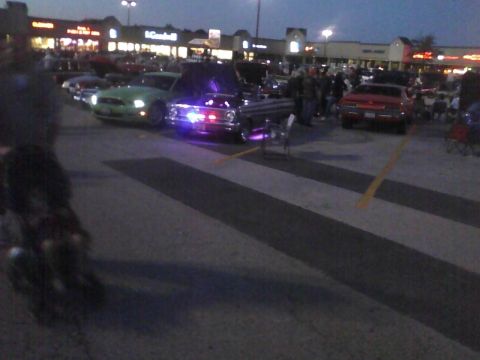 2003 Ford Crown Victoria - Several cars are parked in a lot at dusk, with people milling around and some vehicles displaying lights.