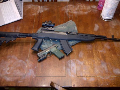1989 Ford Bronco - An SKS rifle with an aftermarket stock and optic, alongside a green pistol, rests on a wooden table.