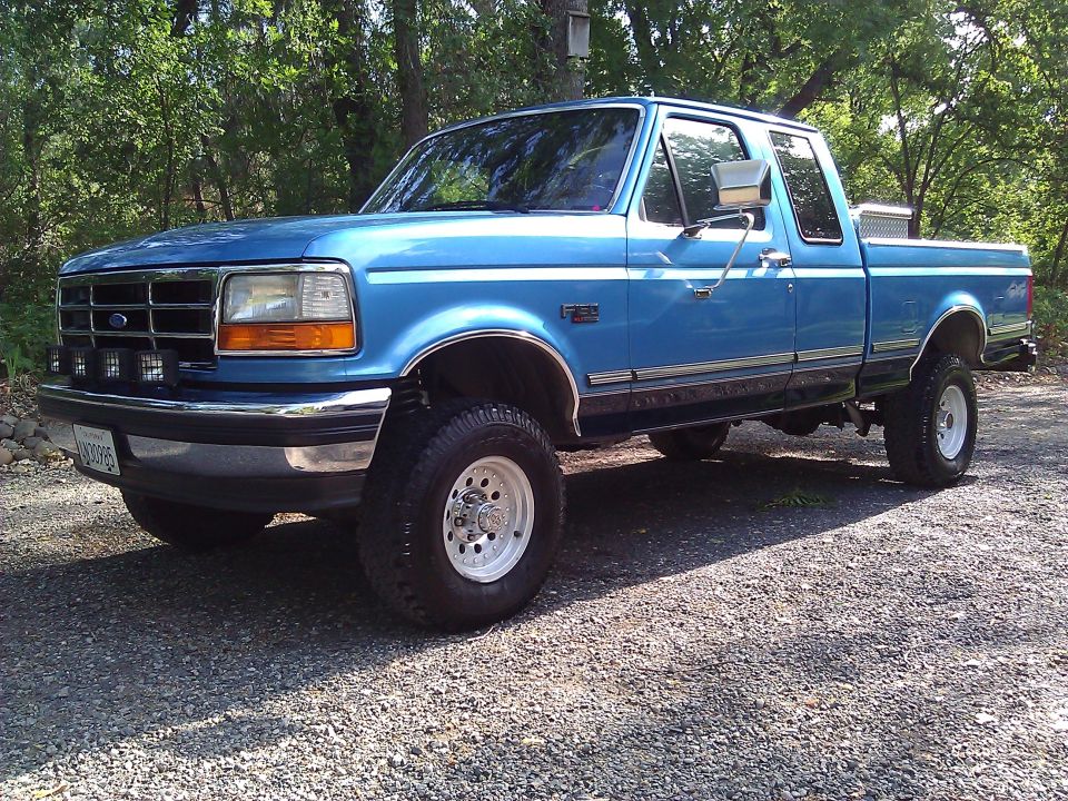 Blue 1992 Ford F-150 pickup truck with chrome accents and aftermarket wheels, parked on gravel