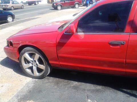 1999 Ford Crown Victoria - A red sedan with custom wheels and a missing headlight is parked on a street.