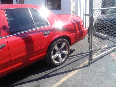 1999 Ford Crown Victoria - Side view of a red sedan with aftermarket wheels, parked near a fence.