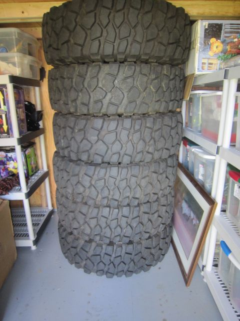 1974 Ford Bronco - Four large, aggressive tread tires are stacked in a storage area next to shelves of plastic bins.
