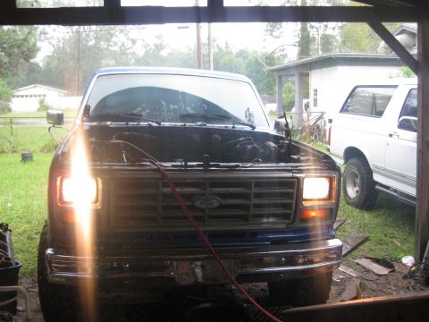 1994 Ford Bronco - Two Ford Broncos are parked in a garage, one with its hood up and headlights on.