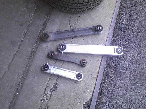 2003 Ford Crown Victoria - A set of four control arms, two aftermarket and two OEM, are displayed on a concrete surface.
