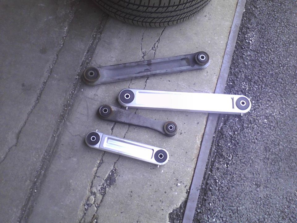 Four car control arms laid out on concrete, one is silver and shiny, three are gray and worn. Tire visible at top