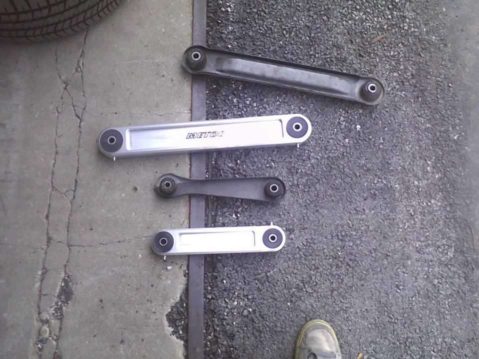 Four control arms laid out on concrete and asphalt, two silver and two black. One silver arm has 'METEOR' text